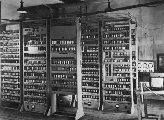 A black and white photograph shows multiple tall vertical racks of the EDSAC computer system densely populated with vacuum tubes and electronic components. The image captures the large-scale physical architecture of early stored-program computers within a laboratory setting.