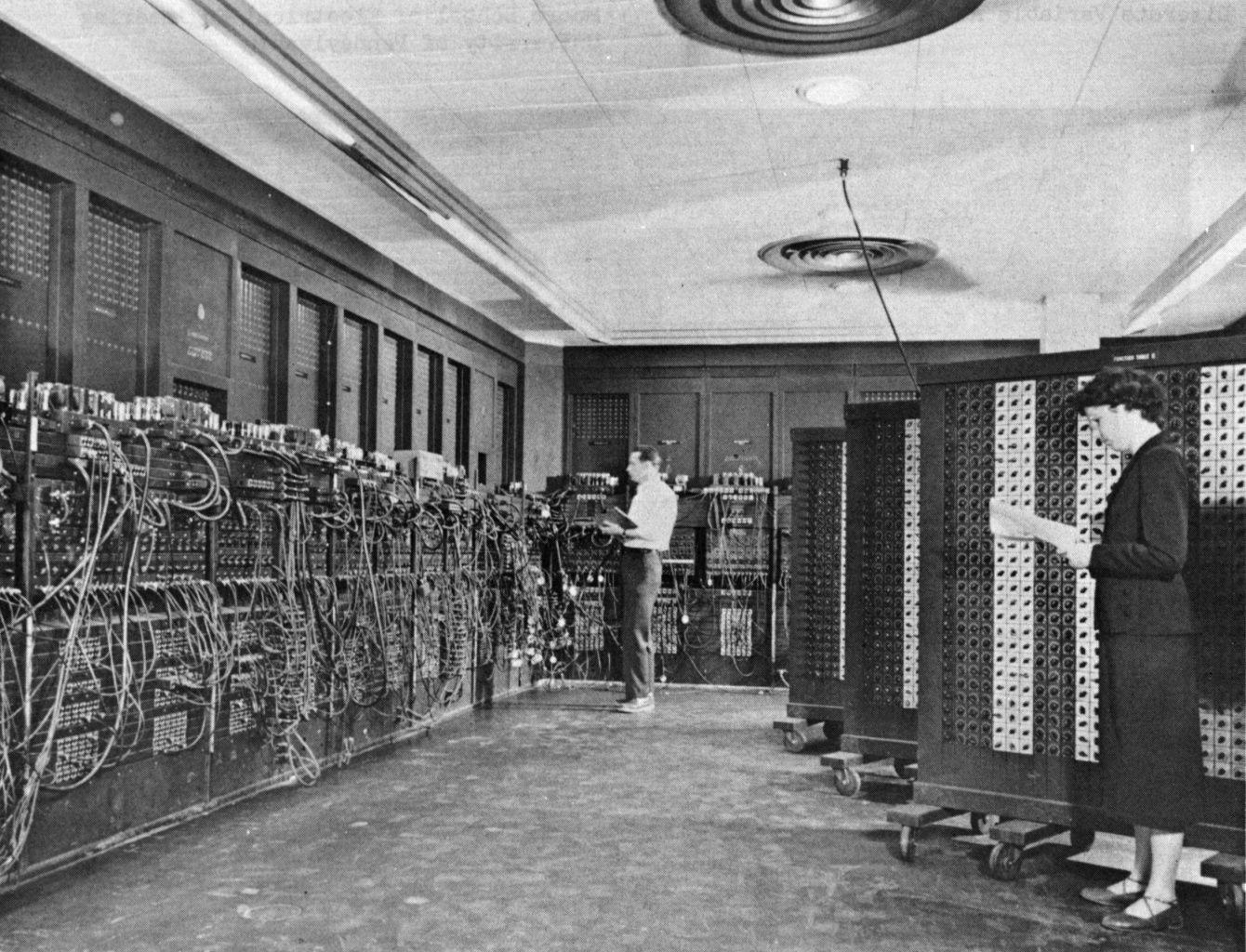 A black and white photograph captures a large room filled with the tall electronic panels and tangled patch cables of the ENIAC computer system. Two people are positioned among the equipment, including a woman in the foreground standing beside a large control cabinet while holding a document.